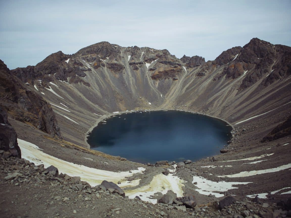 A volcanic crater rim above a deep blue lake, snow patches on the scree