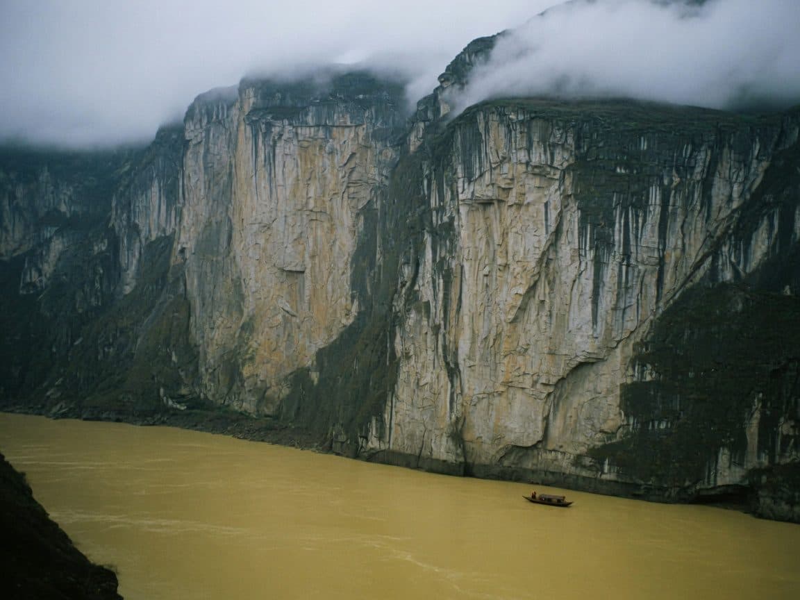 A gorge wall dropping to brown water under low cloud, a small vessel for scale