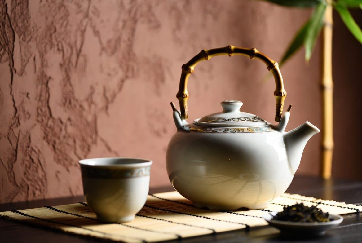 A porcelain teapot, cup, and loose leaf set out on a bamboo mat